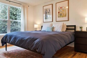 Een slaapkamer met een groot bed, een nachtkastje, een dressoir en twee florale schilderijen in lijsten aan de muur.