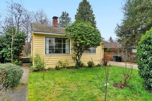 Een gelijkvloers geel huis met voortuin, een kleine boom en een zichtbare schoorsteen.