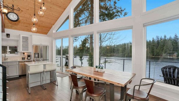 Eine moderne Küche mit großen Fenstern mit Blick auf einen See, ein Essbereich mit einem Holztisch und Metallstühlen sowie ein Ausblick auf den umliegenden Wald.