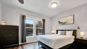 A modern hotel room with a large bed, a nightstand with a lamp, a window with curtains, and a painting on the wall. - New! Prime DT Modern - Extended stay Equipped! (Tacoma)