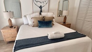 A neatly arranged bedroom with a large bed, two bedside tables, a wall-mounted mirror, and a whale tail artwork.
