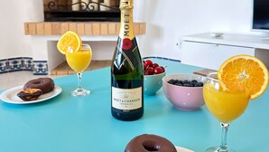 Ruokapöydällä on pullo Moët & Chandon -samppanjaa, lasi appelsiinimehua, suklaadonitsi, portugalilainen munaleivonnainen ja kulho kirsikoita.