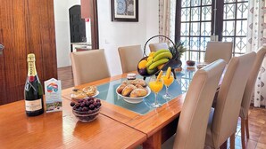 Een eetgedeelte met een houten tafel, stoelen, een fles champagne, een schaal druiven, een fruitmand en een ontbijtbuffet met gebak en sinaasappelsap.