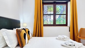 Een hotelkamer met een bed, kussens, een raam met gordijnen, een stoel en een lamp.