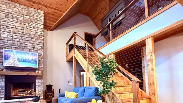 A cozy living room with a fireplace, a blue sofa, a wooden staircase, and a stone wall. - PoStickNo Lodge: 6BR/6.5BA Ski-In/Out Pet-Friendly (Snowshoe)