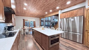 A modern kitchen with wooden cabinetry, a central island, stainless steel appliances, and a view of a forested area. - PoStickNo Lodge: 6BR/6.5BA Ski-In/Out Pet-Friendly (Snowshoe)