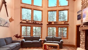 A spacious living room with a high ceiling, large windows, a fireplace, and a coffee table with a book. - PoStickNo Lodge: 6BR/6.5BA Ski-In/Out Pet-Friendly (Snowshoe)