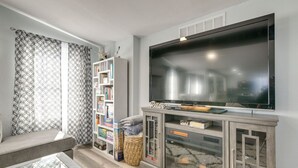 A living room with a large flat-screen TV, a bookshelf, and a window with patterned curtains. - Galveston Beach Retreat | Balcony & Walk to Seawall (Galveston)
