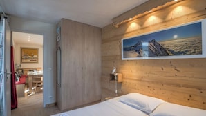 Quarto com parede de madeira, cama com roupa de cama branca, quadro pendurado de uma montanha e porta de madeira que dá para uma sala de jantar.