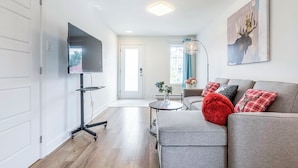 A modern living room with a grey sofa, red and plaid cushions, a flat-screen TV mounted on a stand, a floor lamp, and a painting of a deer on the wall. - 10-Min La Ronde Ride! Cozy Place, Free Parking! (Longueuil)