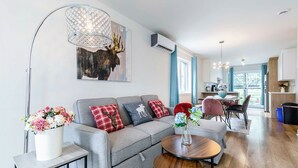 A modern living room with a grey sofa, a round wooden coffee table, and a floor lamp. There is a painting of a moose on the wall, and a kitchen area is visible in the background. - 10-Min La Ronde Ride! Cozy Place, Free Parking! (Longueuil)