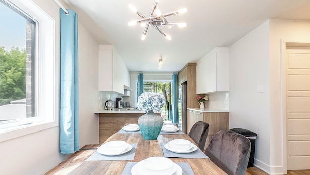 A modern kitchen with white cabinets, a wooden dining table set with plates, and a decorative vase. - 10-Min La Ronde Ride! Cozy Place, Free Parking! (Longueuil)