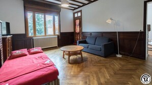 A room with a wooden floor, a sofa, a table, a TV, and a window with a view of trees. - Chez Marius - ESTENOS (ESTENOS)