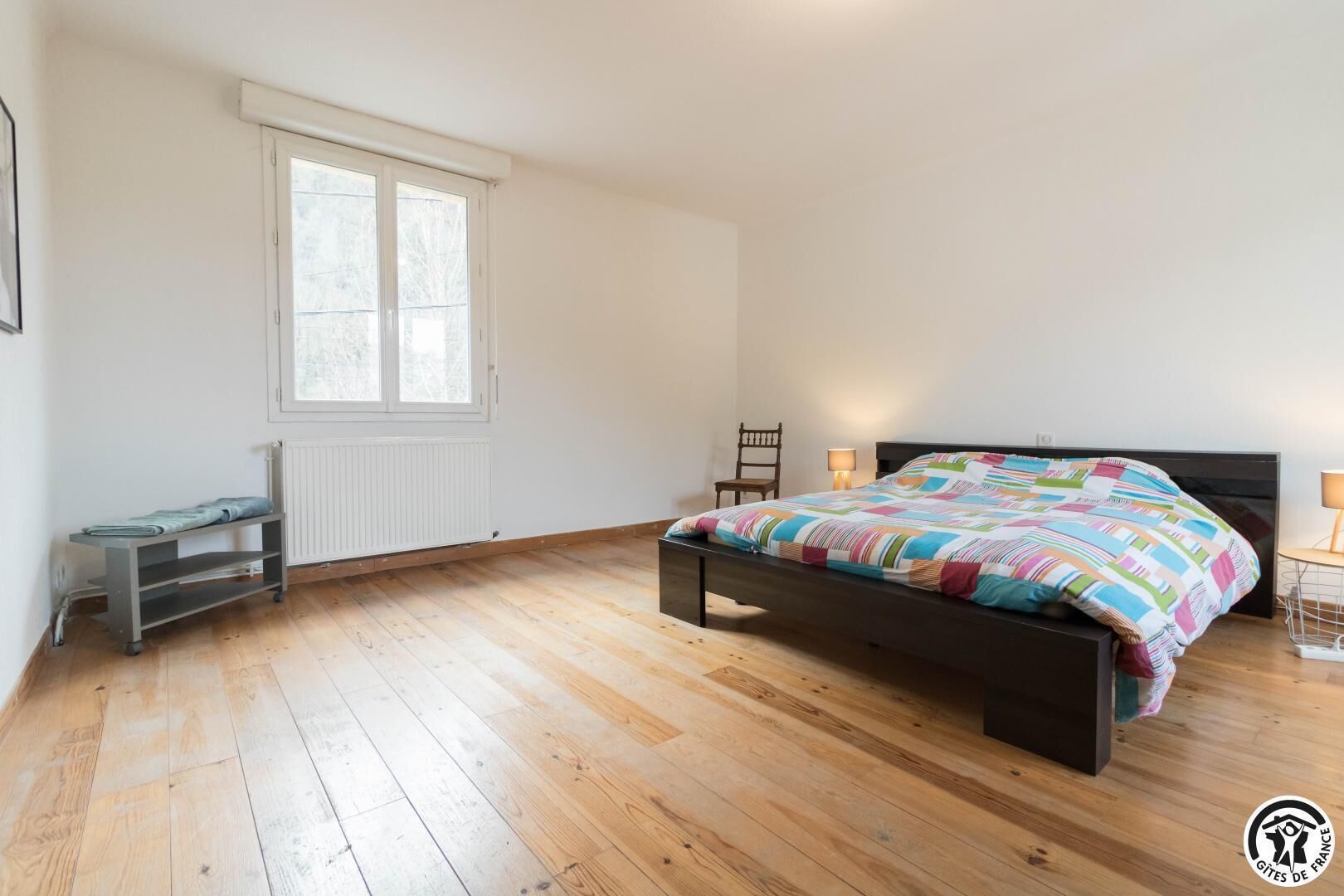 Une chambre avec un plancher en bois, un lit recouvert d’une couverture aux couleurs vives, une chaise, un bureau et une fenêtre.