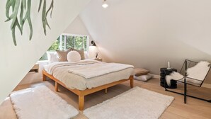 A modern bedroom with a sloped ceiling, a wooden bed, white fluffy rugs, and a black chair.
