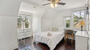 Un dormitorio con cama, escritorio, silla, ventilador de techo y ventanas grandes.