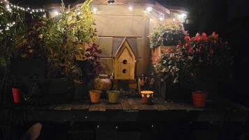Un entorno rústico al aire libre con un comedero de pájaros de madera, plantas en macetas y luces de cuerda.