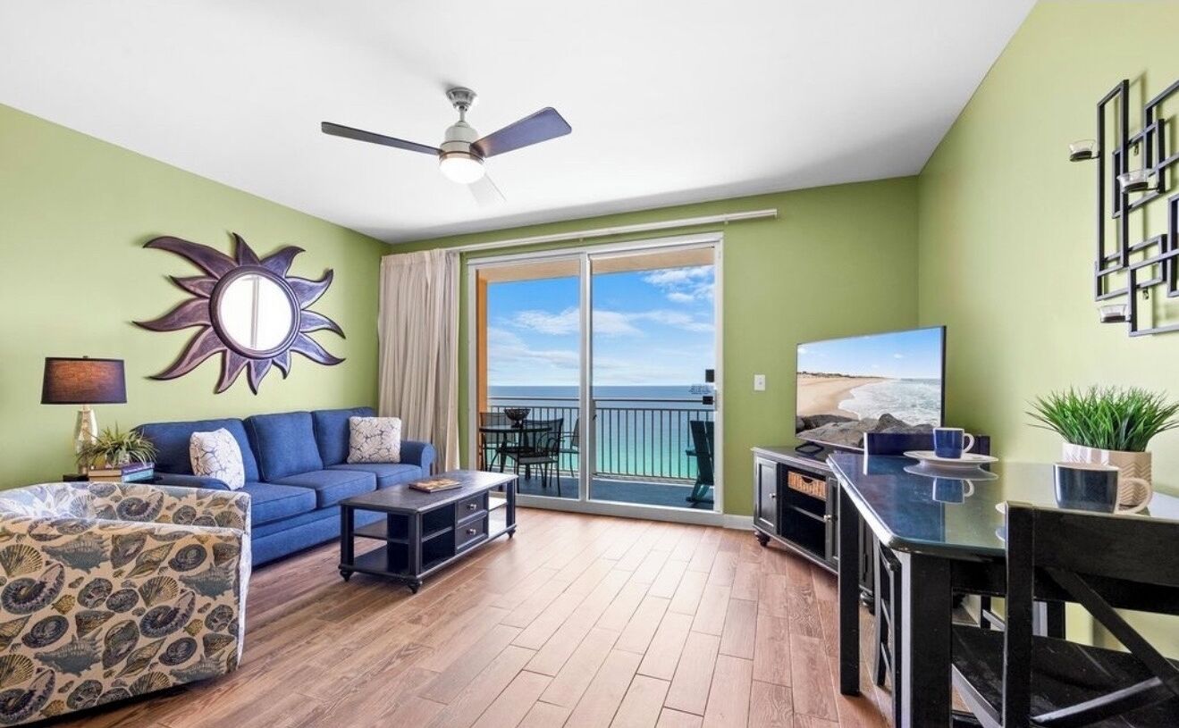 Ein Wohnzimmer mit einem blauen Sofa, einem gemusterten Hocker, einem Couchtisch und einem Fernseher. Ein Balkon bietet Blick aufs Meer.