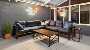Un'area salotto esterna moderna con un ampio divano componibile, un tavolino in legno e un piccolo tavolino laterale. Un neon recita "The Mango-Tree House".
