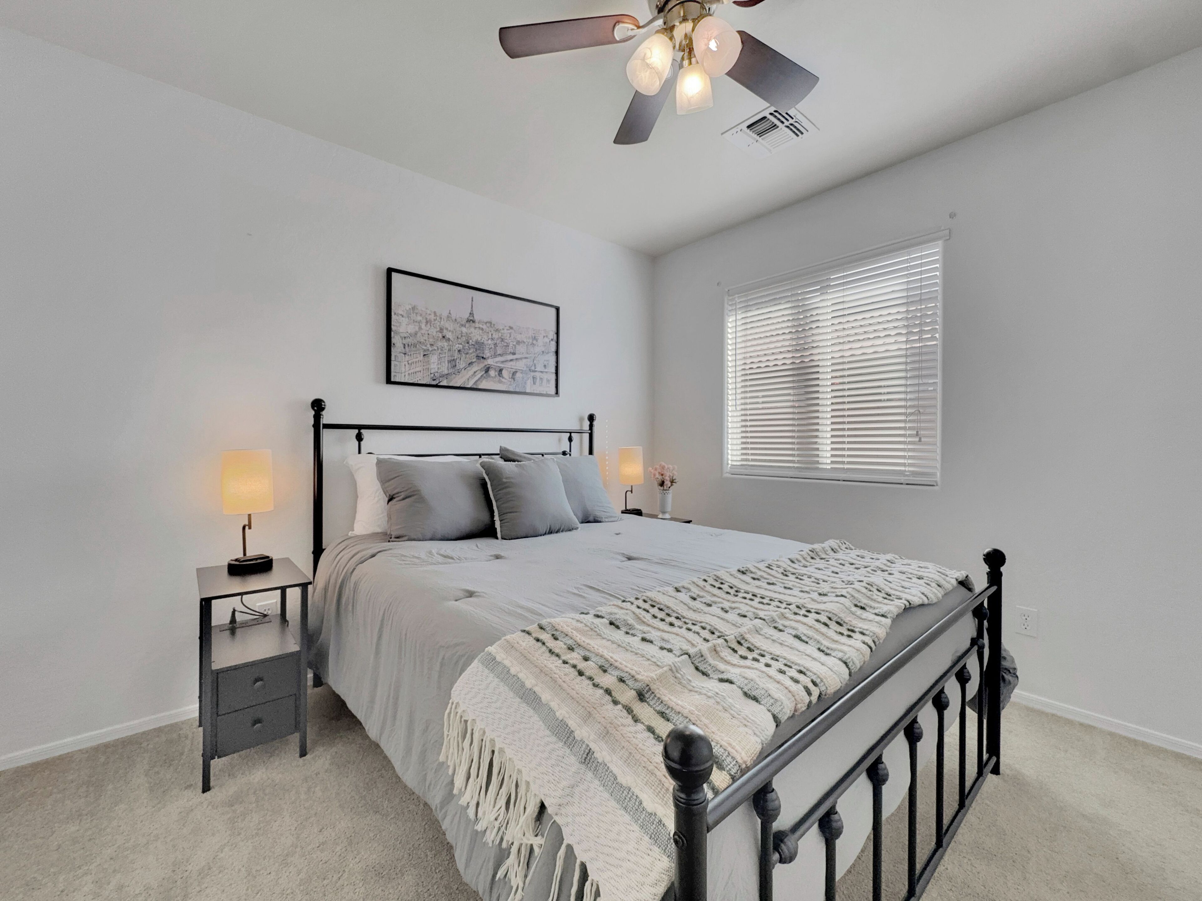 Quarto com cama, mesa de cabeceira, ventilador de teto, janela com persianas e um quadro na parede.