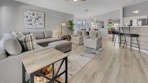 A spacious living area with a sofa, ottoman, and a dining table. There is a ceiling fan, a chandelier, and a kitchen area visible in the background. - Single-Level 4BR Surprise Home | Heated Pool, Game Room, Arcade (Surprise)