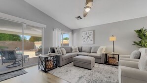 A spacious living room with a grey sofa, a white armchair, and a coffee table. There is a large window with a view of the outside, and a ceiling fan is visible. - Single-Level 4BR Surprise Home | Heated Pool, Game Room, Arcade (Surprise)