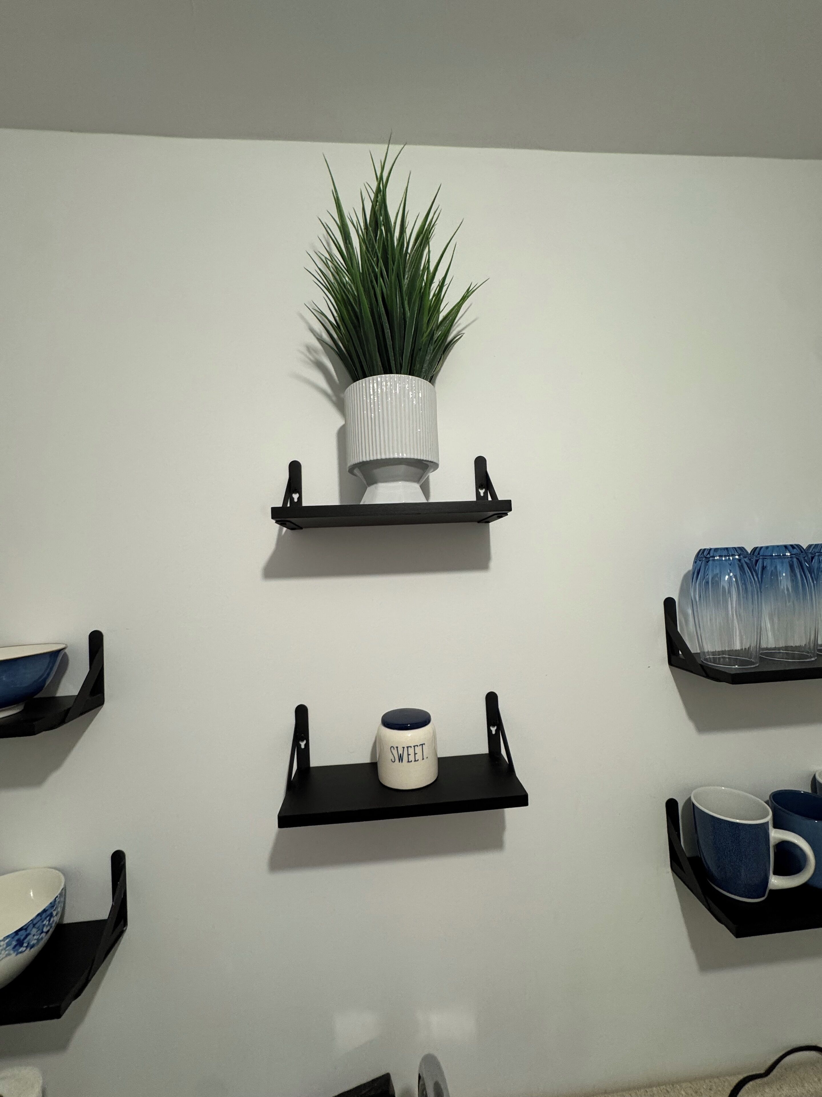 Un mur orné de trois étagères noires, chacune contenant des objets différents : une plante en pot, une tasse et un ensemble de verres.