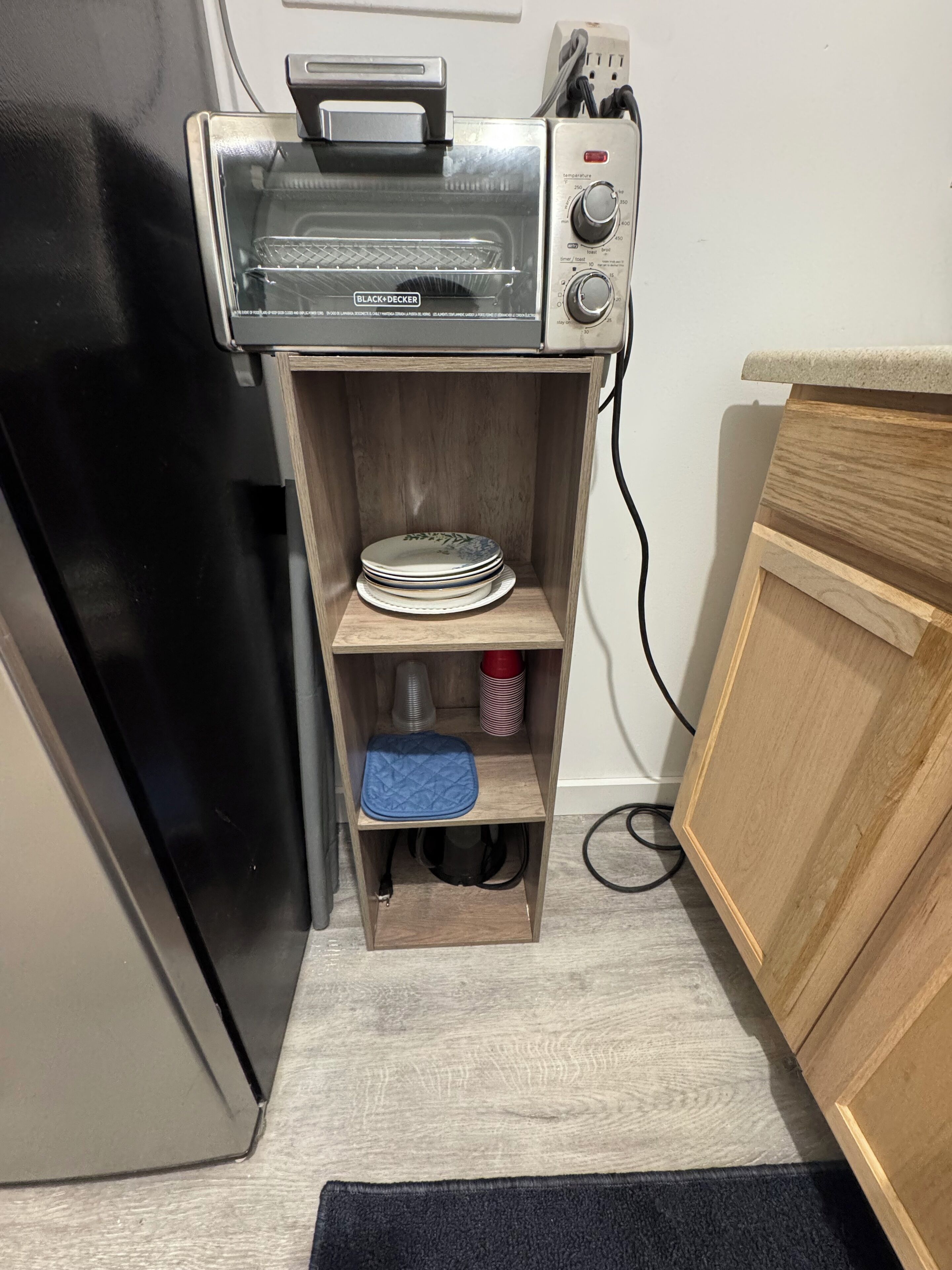 A kitchen with a built-in toaster oven, a wooden shelf with plates and a blue cloth, and a black refrigerator.