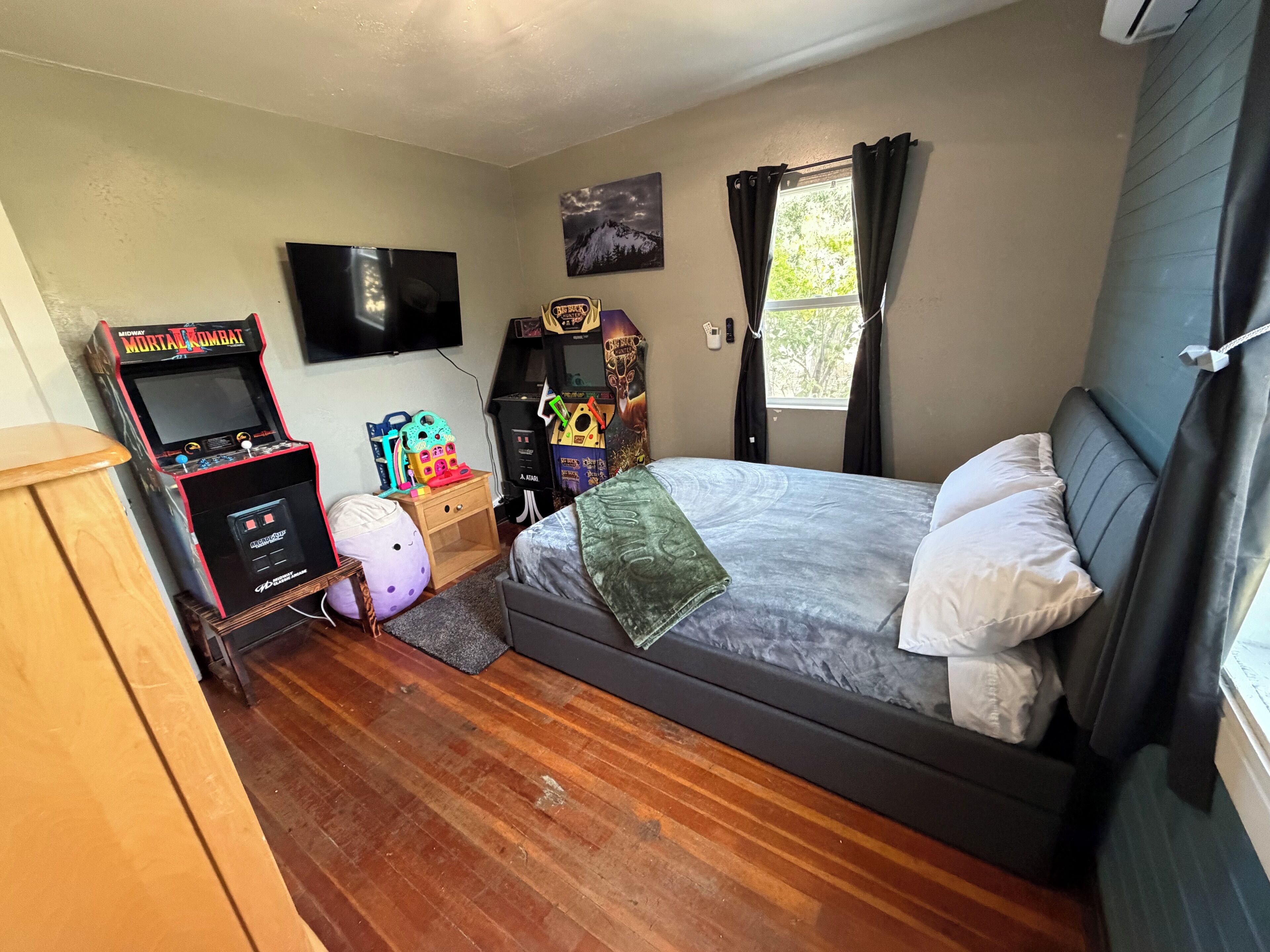 A bedroom with a bed, a television, a gaming console, and a window with curtains.