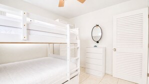 Una cama litera con cabecera blanca, un aparador con espejo y una puerta blanca con persianas horizontales.