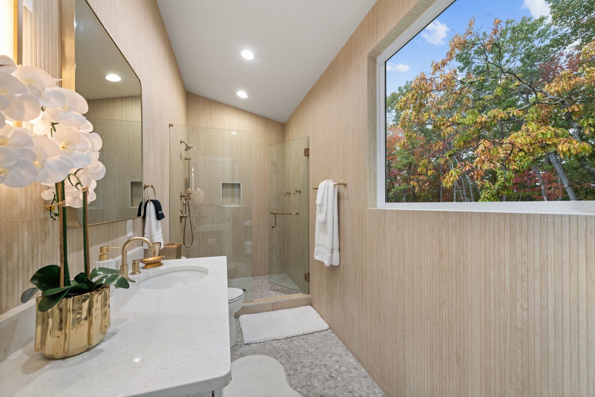 A modern bathroom with a large mirror, a white sink, a shower, and a view of autumn trees.