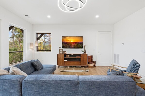 A modern living room with a large sofa, a wooden cabinet, and a painting of a sunset. - Blue Ridge Mountain Retreat, Panoramic Views, Game Room, and Hot Tub! (Blue Ridge)