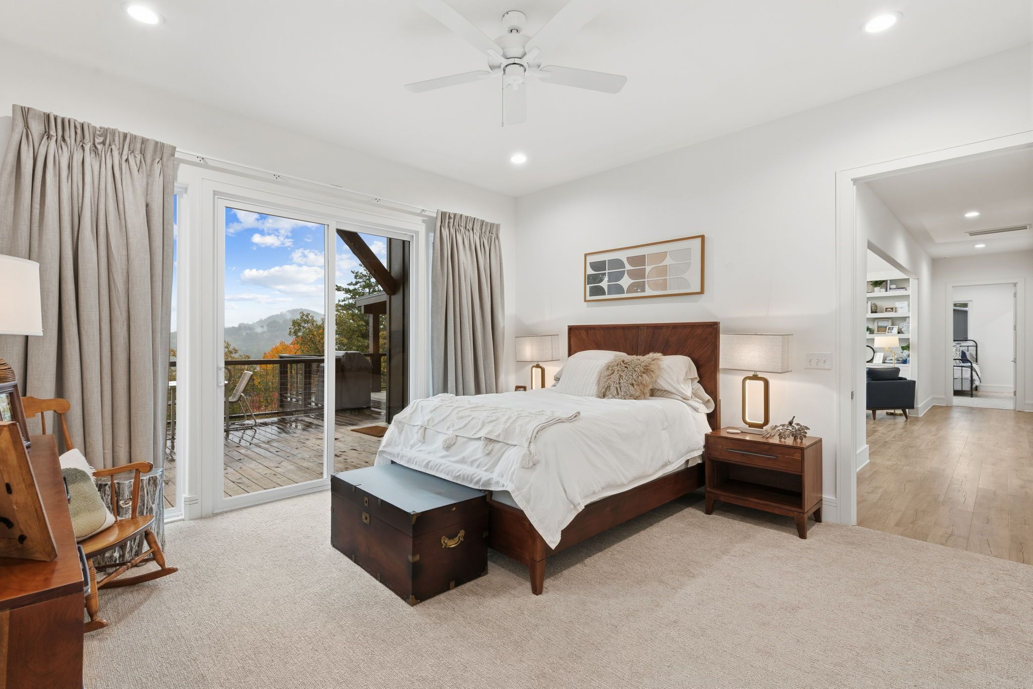 Une chambre avec un grand lit, une table de chevet en bois, une commode, un fauteuil à bascule et une vue sur l’extérieur grâce à une porte-fenêtre coulissante.