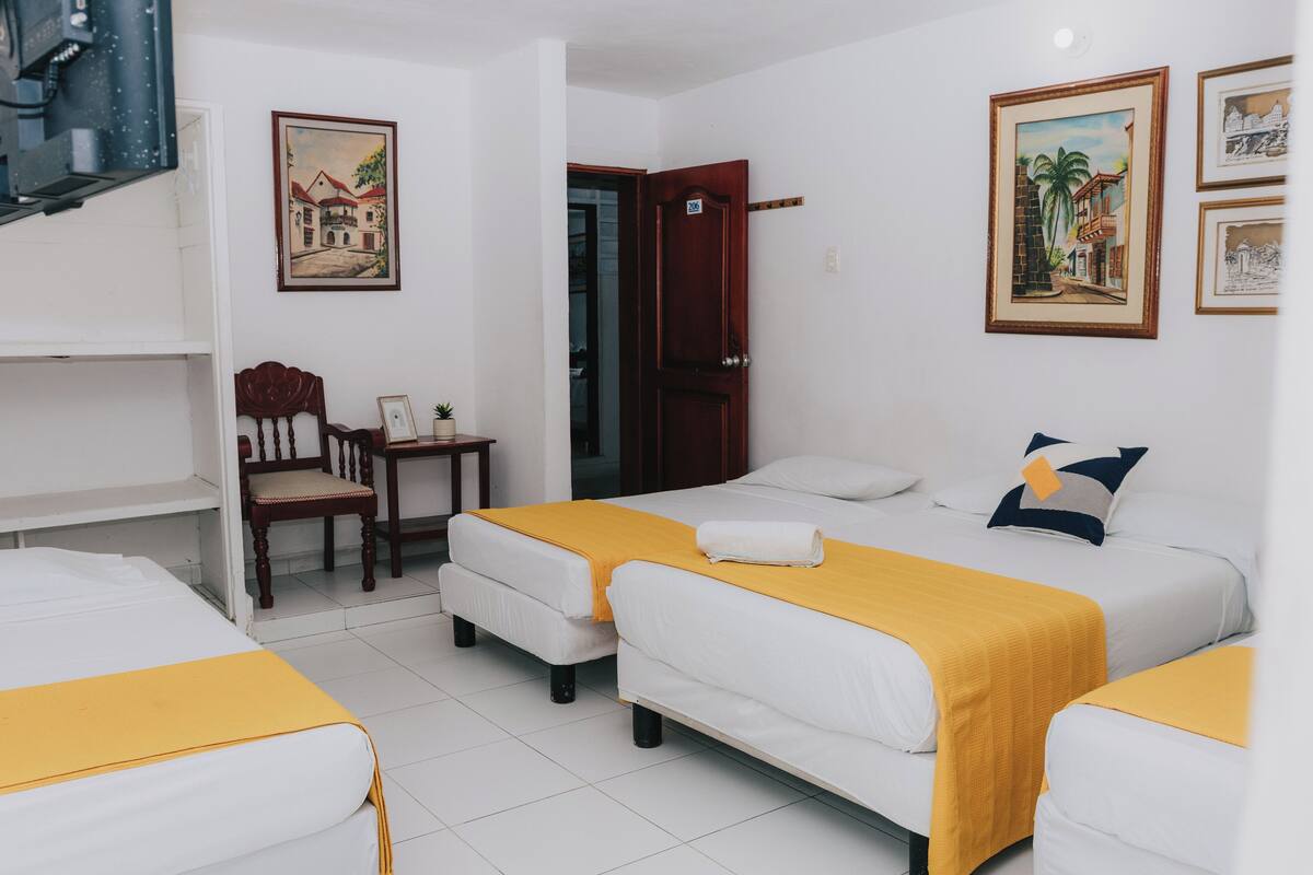 Habitación de hotel con dos camas, una silla de madera, una mesita y una puerta que da a otra habitación.
