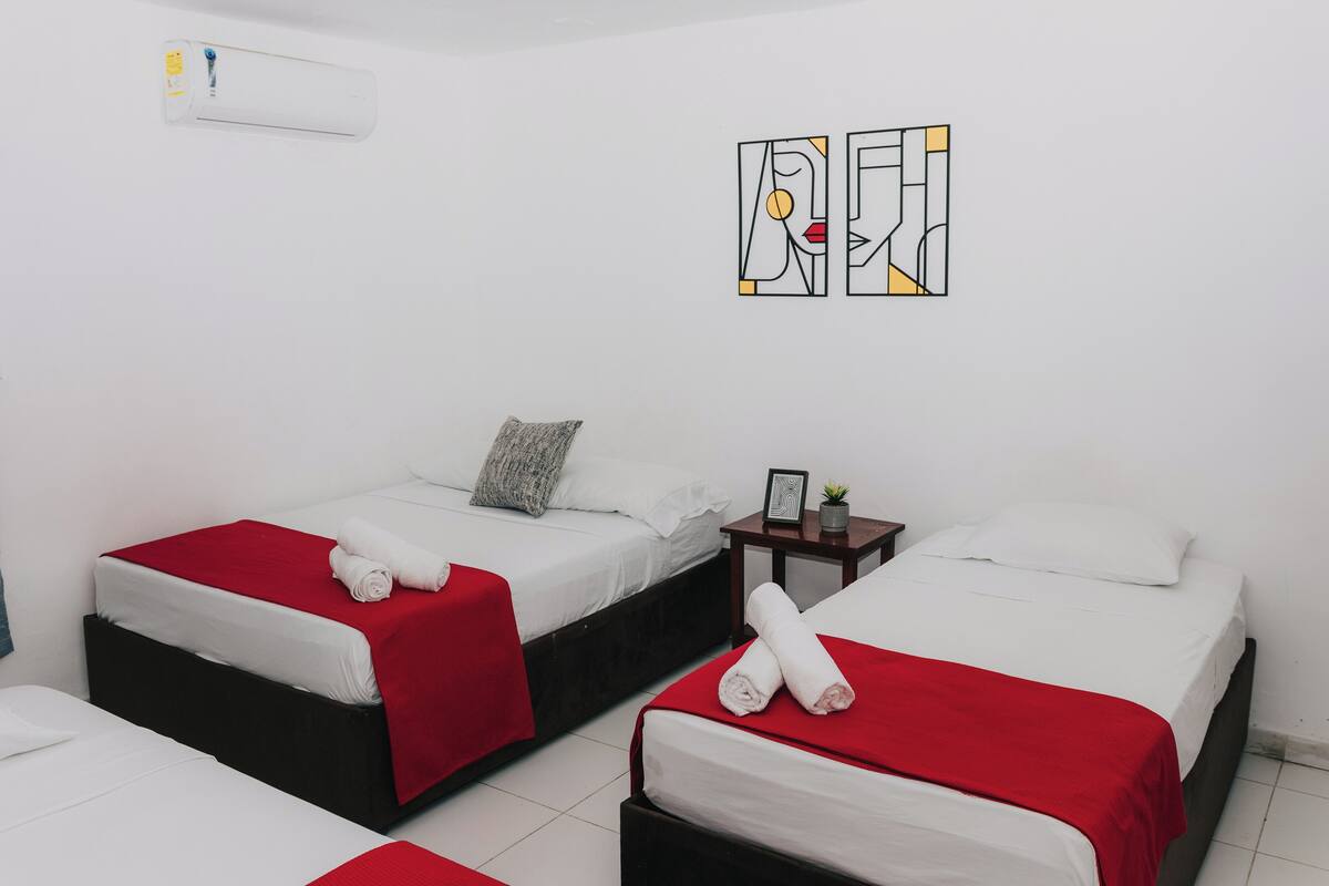 Habitación de hotel con dos camas, cada una con cubrecama rojo y ropa de cama blanca. Se observa un pequeño tocador de madera con un cuadro enmarcado y una planta.