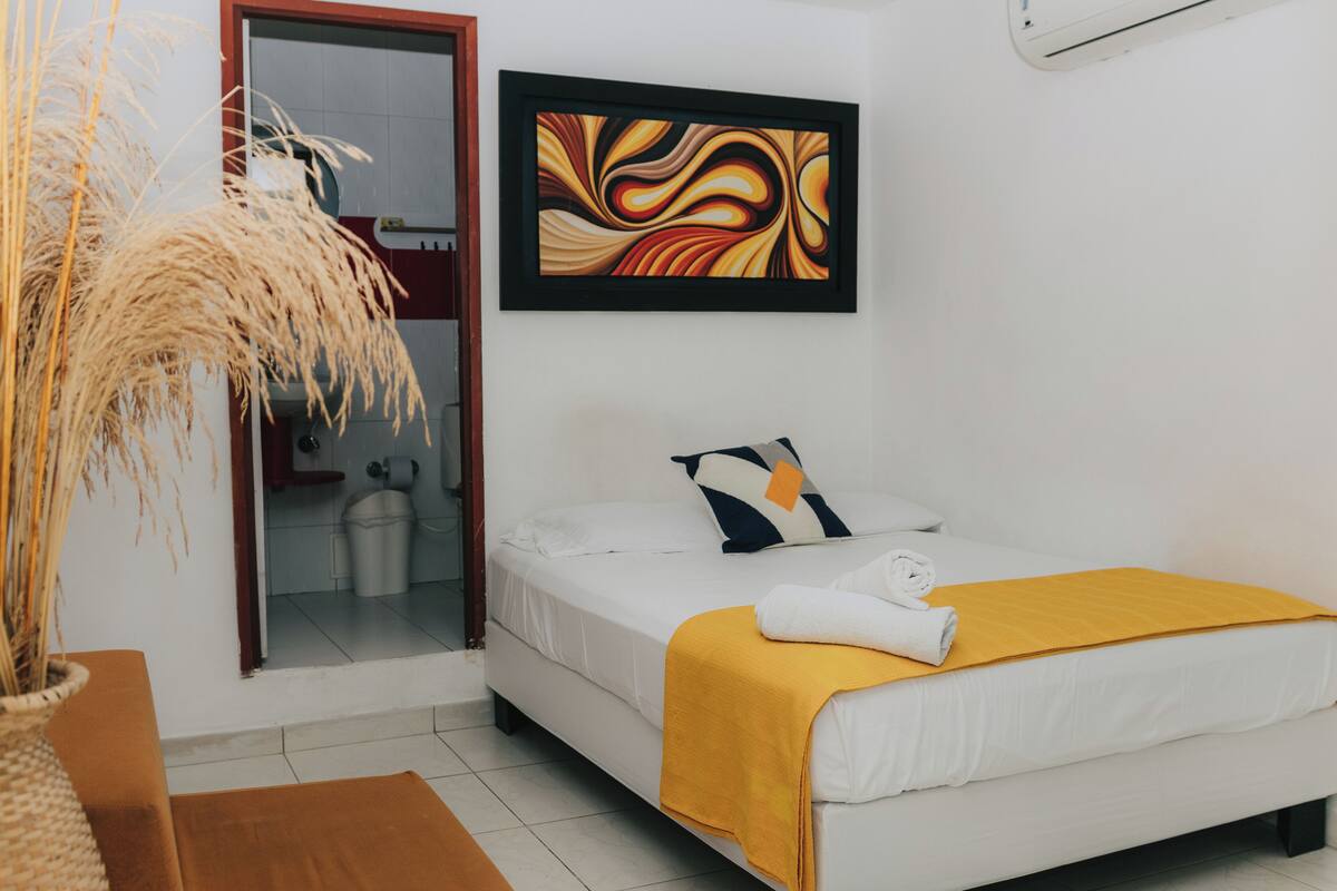Habitación de hotel con una cama, una manta amarilla, un almohadón, un cuadro abstracto enmarco y una planta en maceta.