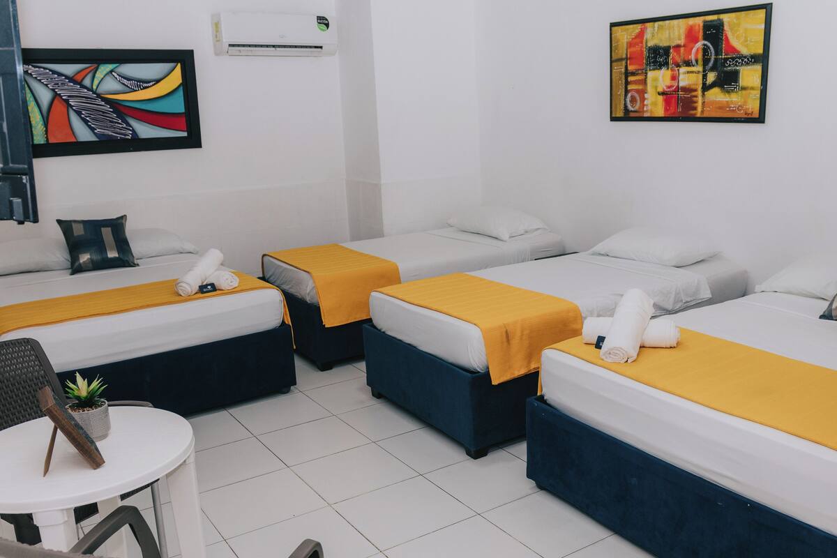 Habitación de hotel con tres camas, cada una con una manta amarilla y almohadas blancas. Hay una mesita con una planta y una silla.