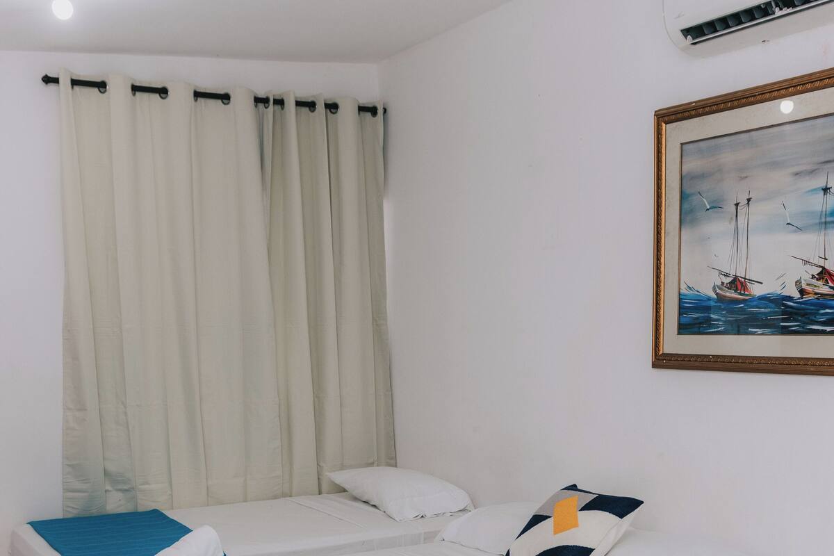 Habitación de hotel con dos camas individuales, un ventanal con cortinas, un cuadro en la pared y un equipo de aire acondicionado.
