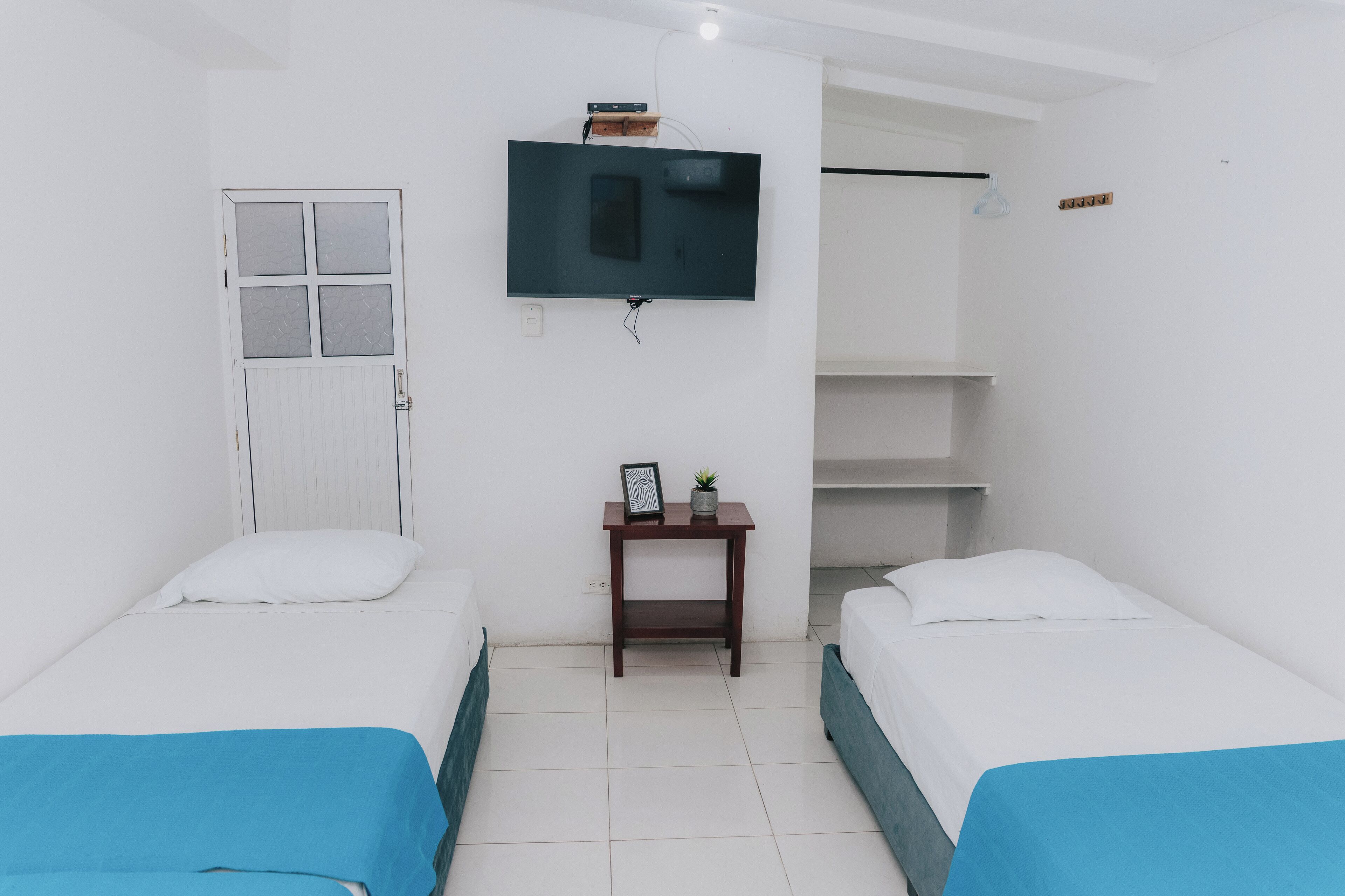 A room with two beds, a TV mounted on the wall, a small table with a plant, and a window with a door.