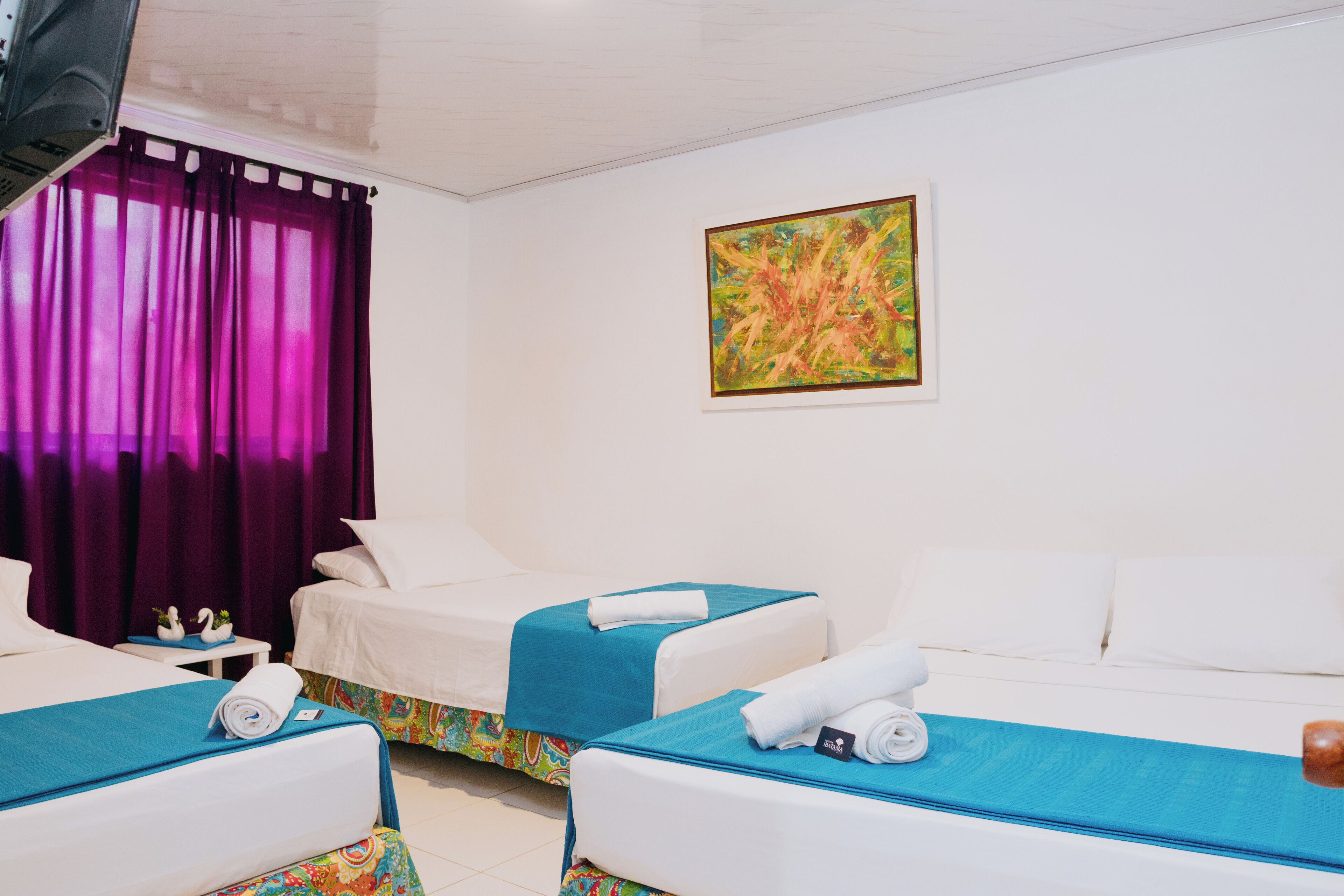 Habitación de hotel con dos camas, una colcha colorida, un cuadro en la pared y cortinas moradas.