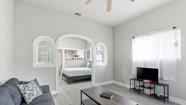 Una sala moderna con un sofá gris, una mesa de centro, un televisor sobre un mueble bajo, y un dormitorio con una cama y ventanas arqueadas.