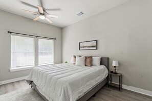 Un dormitorio con cama, ventilador de techo, mesita de noche, lámpara y ventana con persianas.