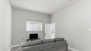 A living room with a grey sofa, a television on a stand, and a window with blinds. - Bright 2BR Duplex Mins to Downtown San Antonio (San Antonio)