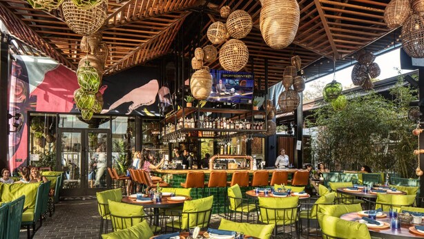 Un restaurant en plein air avec des tables rondes et des chaises vertes, un bar avec des étagères remplies de bouteilles et des lampes en osier suspendues.