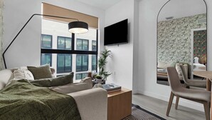 Ein modernes Hotelzimmer mit großem Fenster, einem Flachbildfernseher, einem Schreibtisch mit Stuhl und einem Bett mit grüner Decke.