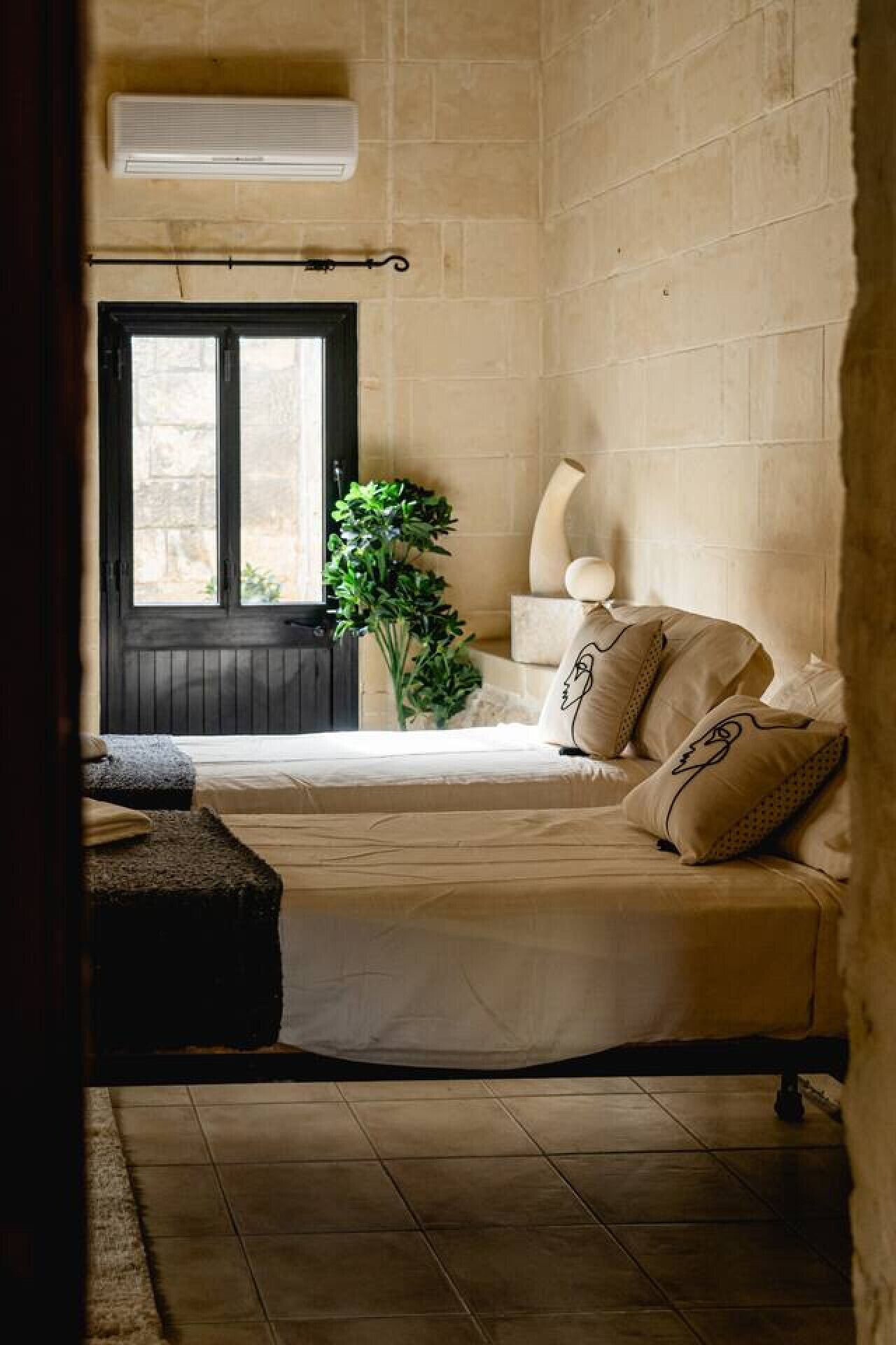 A cozy room with a bed, a window, a potted plant, and a wall-mounted air conditioner.