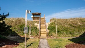 Een houten trap die leidt naar een houten gebouw met een dak, omringd door gras en duinen.
