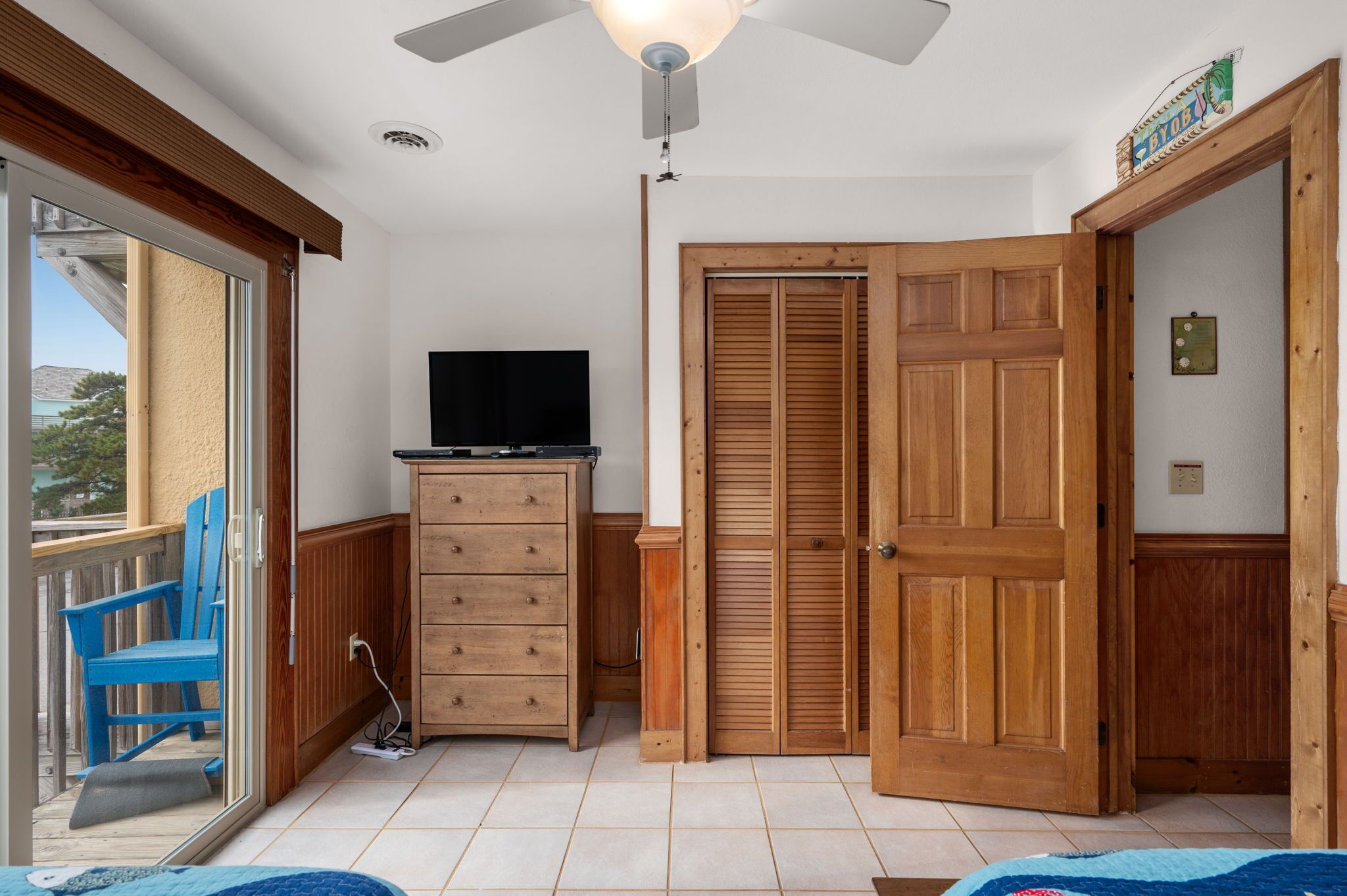A room with a sliding glass door leading to a balcony, a wooden cabinet with a television, and a wooden door with louvered panels.