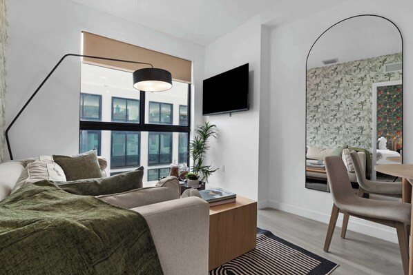 Una habitación de hotel moderna con un ventanal grande, un televisor de pantalla plana, un escritorio con una silla y una cama con manta verde.