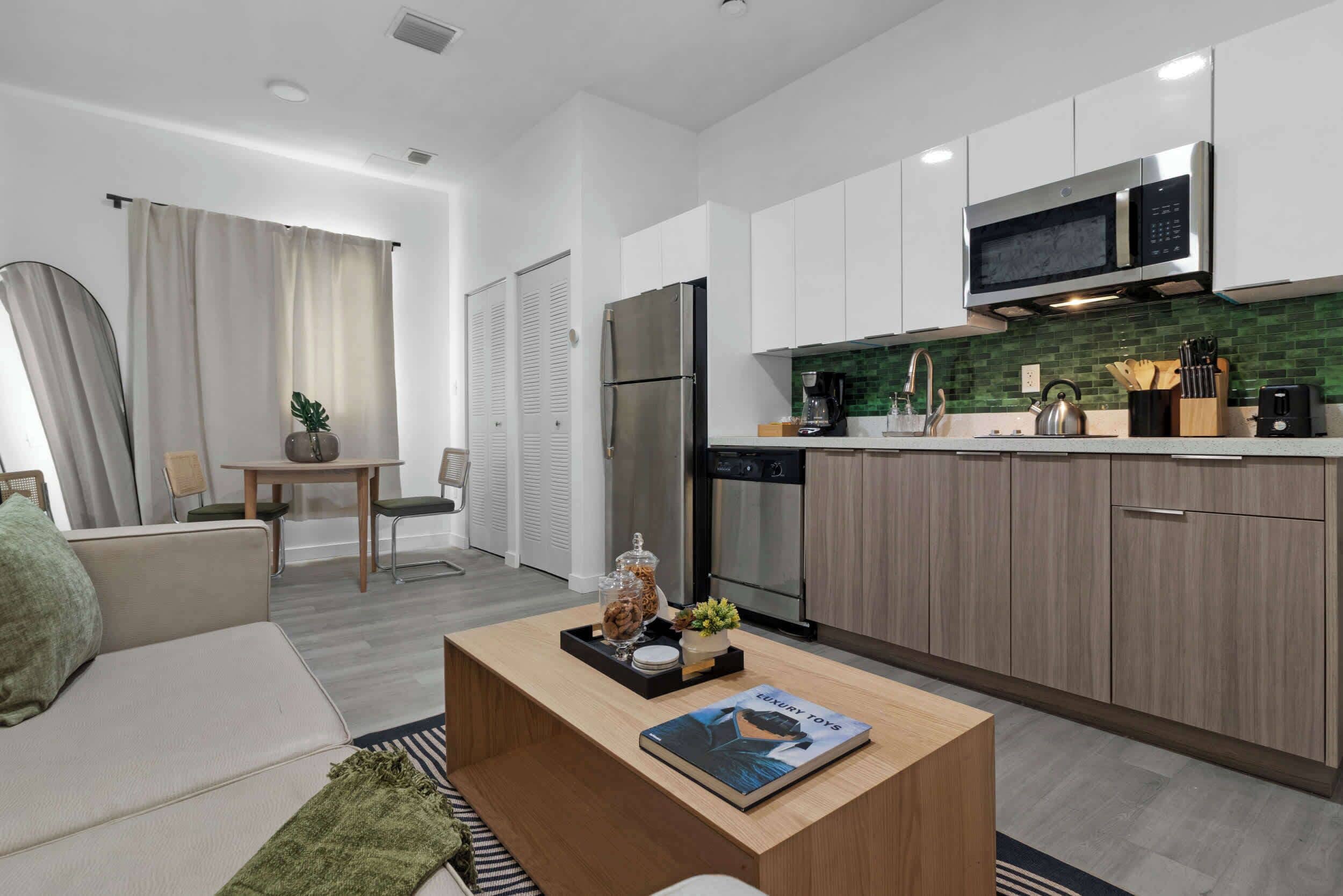 Eine moderne Küche mit weißen Schränken, einem Edelstahlkühlschrank und einer Mikrowelle. Es gibt einen Essbereich mit Tisch und Stühlen sowie ein Wohnzimmer mit Sofa und Couchtisch.
